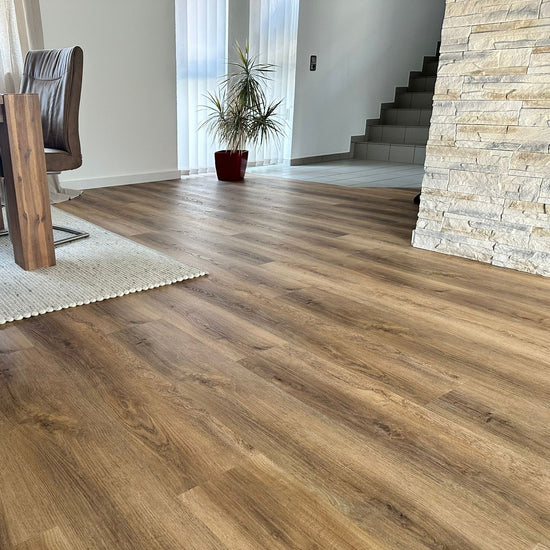 Eiche Eiche Apatura Klebevinyl in Wohnzimmer mit Holztisch, Stühlen, Teppich und Steinmauer